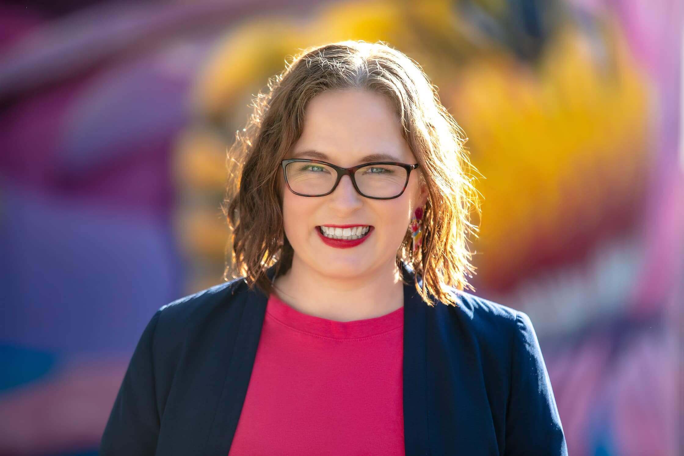 A woman with brown hair glasses  is wearing a navy jacket and pink top. No Grey Suits talks about service packaging.